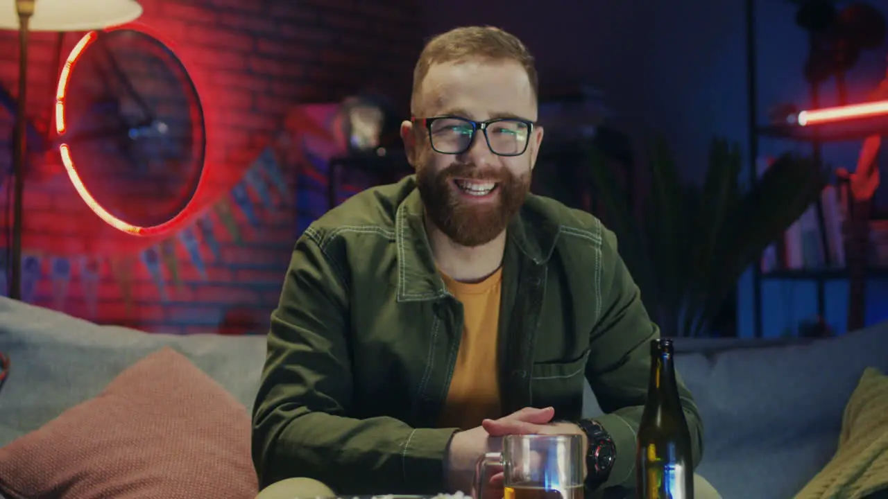 Portrait Of The Joyful Red Haired Young Man In Glasses Smiling To The Camera And Being In Good Mood While Sitting On The Sofa In The Dark Room