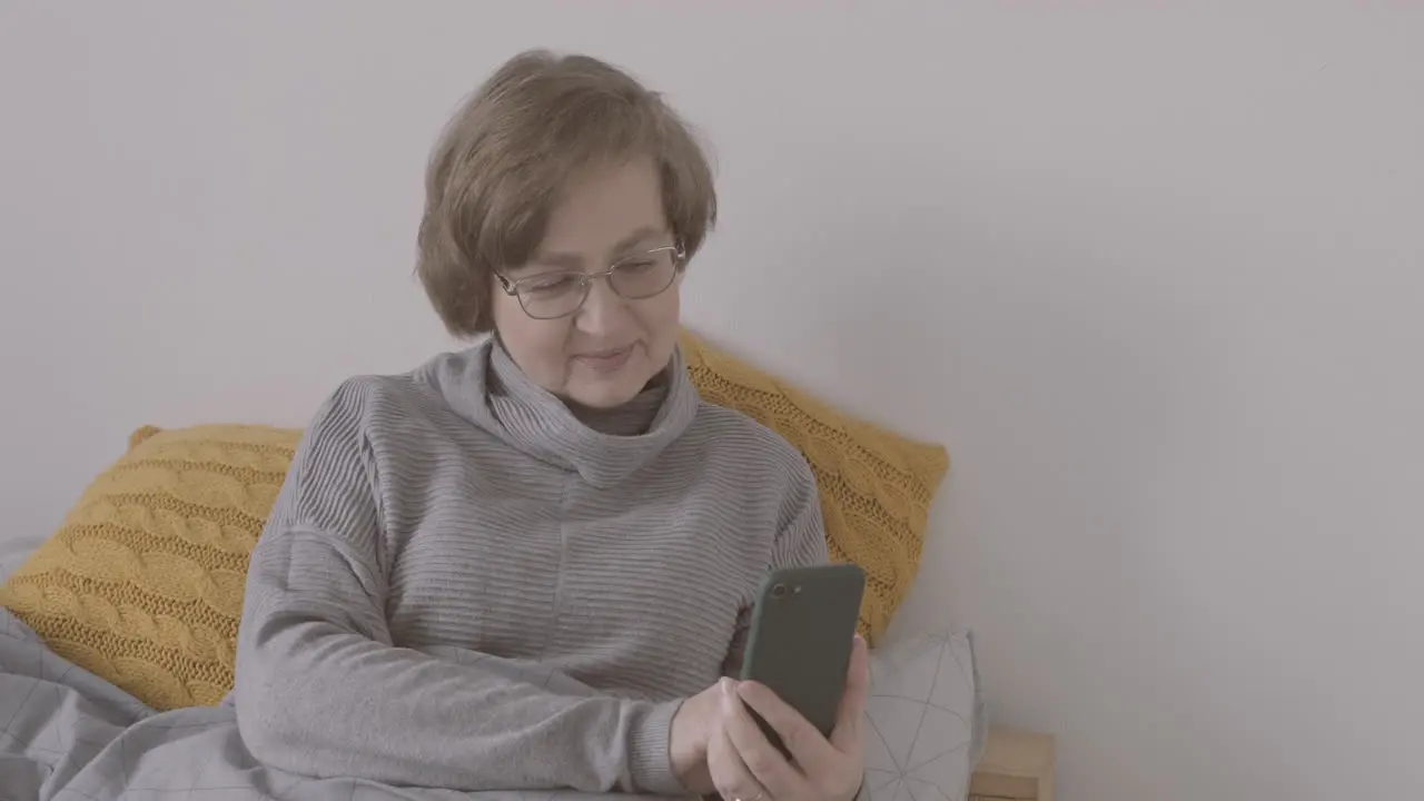 Senior Woman With Eyeglasses Sitting On Bed Using Smartphone