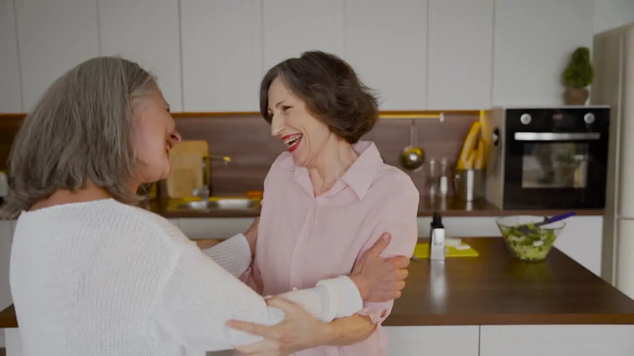 Senior Woman Waving And Hugging Her Older Friend In The Kitchen