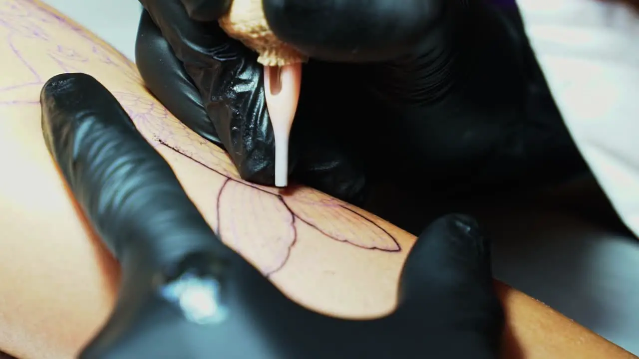 Close up slow motion handheld shot of the needle of a tattoo artist penetrating the skin as they follow the temp outline of the desired image in a dark studio room