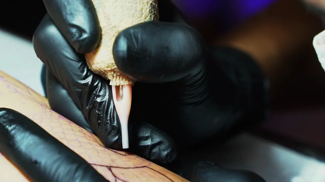 Close up slow motion tilt down shot of the needle of a tattoo artist penetrating the skin as they follow the temp outline of the desired image in a dark studio room