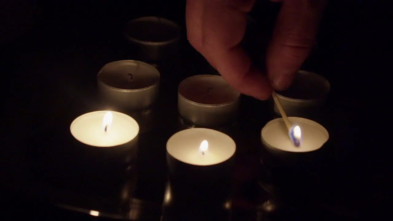 Fingers hold burning wooden match to light tea candles in dark room