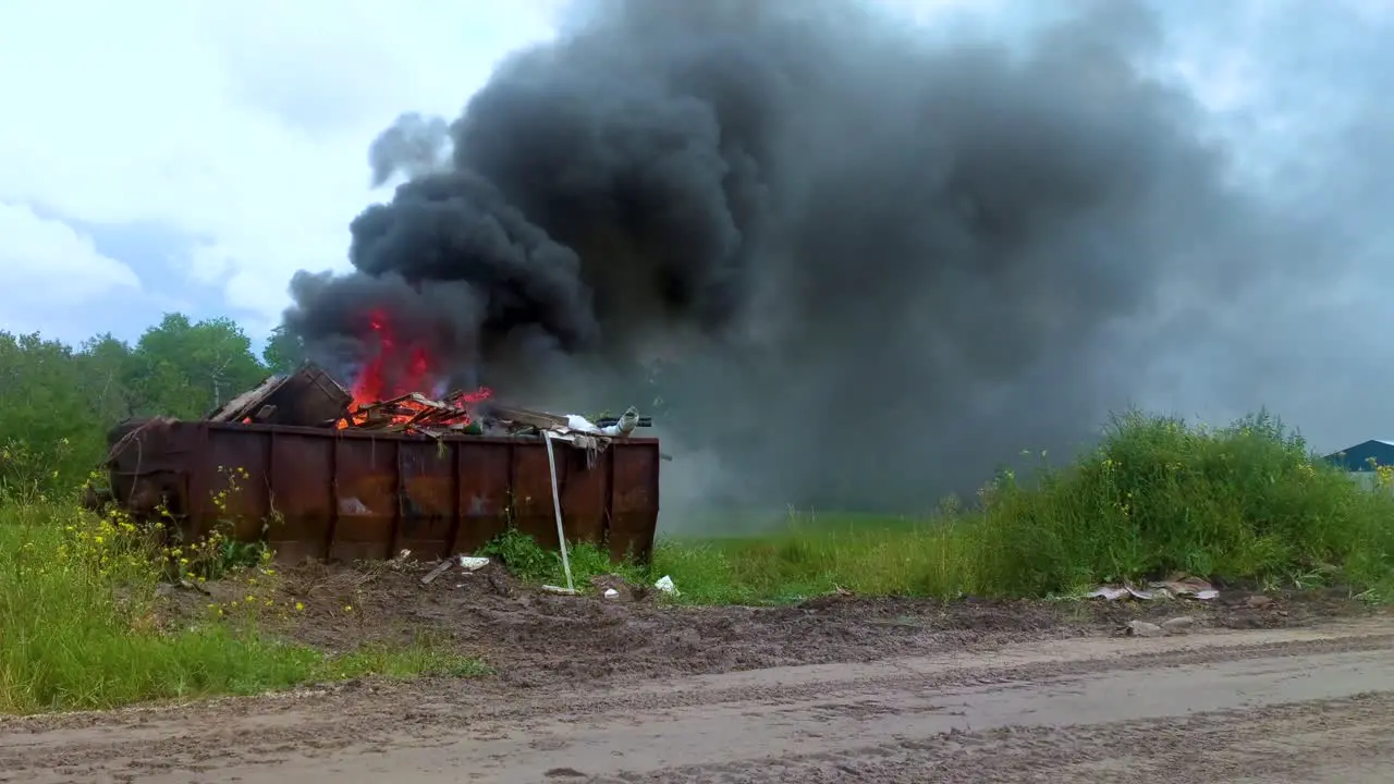 Thick dark smoke billowing from a burning dumpster full of garbage on rural farmland