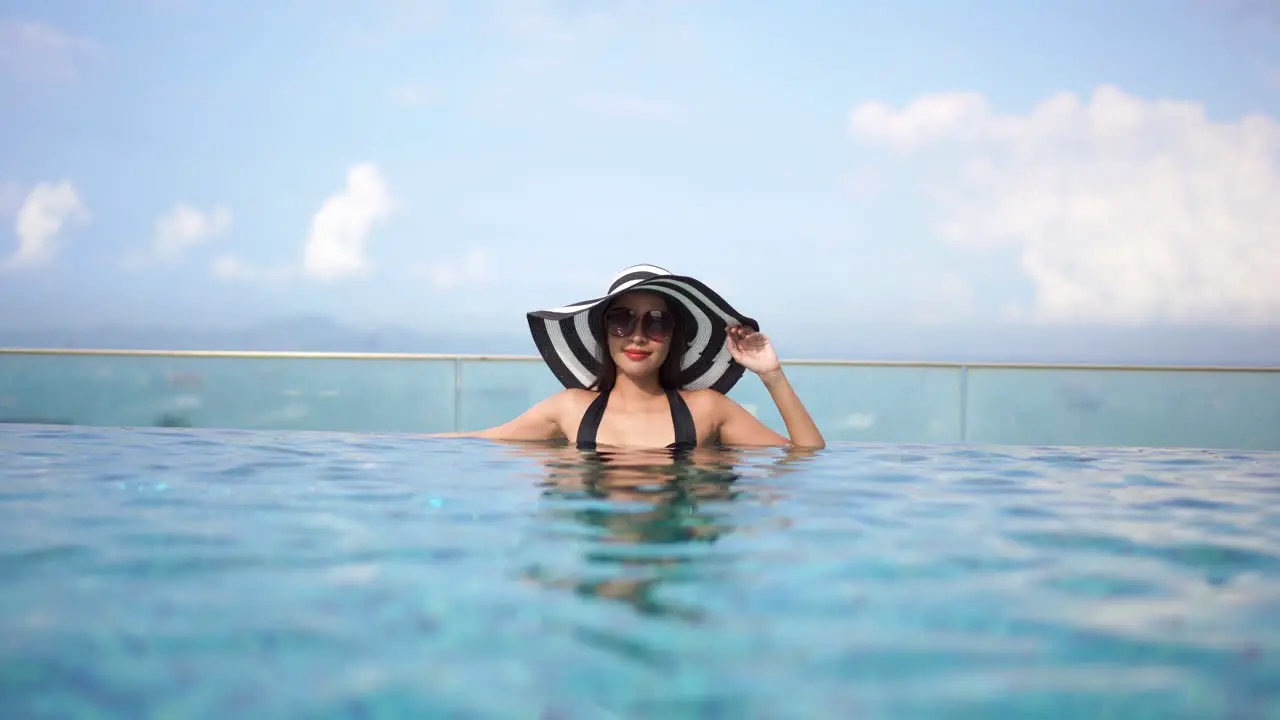 Beautiful woman with a pretty smile wearing a sun hat and glasses relaxes in the clear rippling waters of a lavish infinity pool under bright blue skies with white clouds and the sea in the background