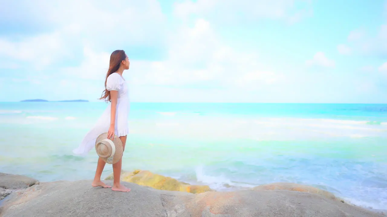 Woman with hat in hand and dressed in white admiring blue sea from cliff