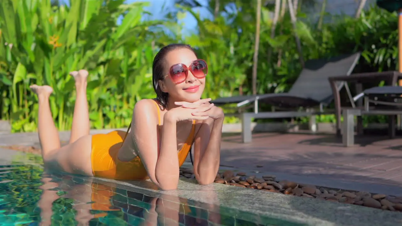 Attractive hot Asian Woman Lying on her Belly on the Border of the Swimming Pool of Tropical Hotel Looking Straight Into the Camera Lens Smiling While Moving Legs Up and Down slow motion