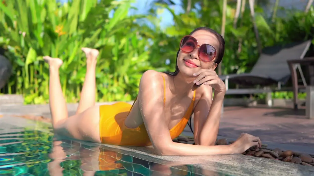 Smiling Asian girl with big sunglasses sunbathes lying on edge of pool