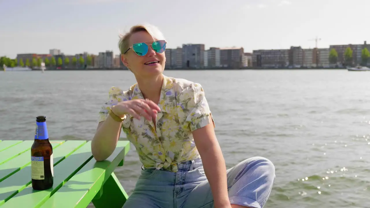 Fun young woman enjoys the summer sun with a beer sitting on picnic table river and city in the background