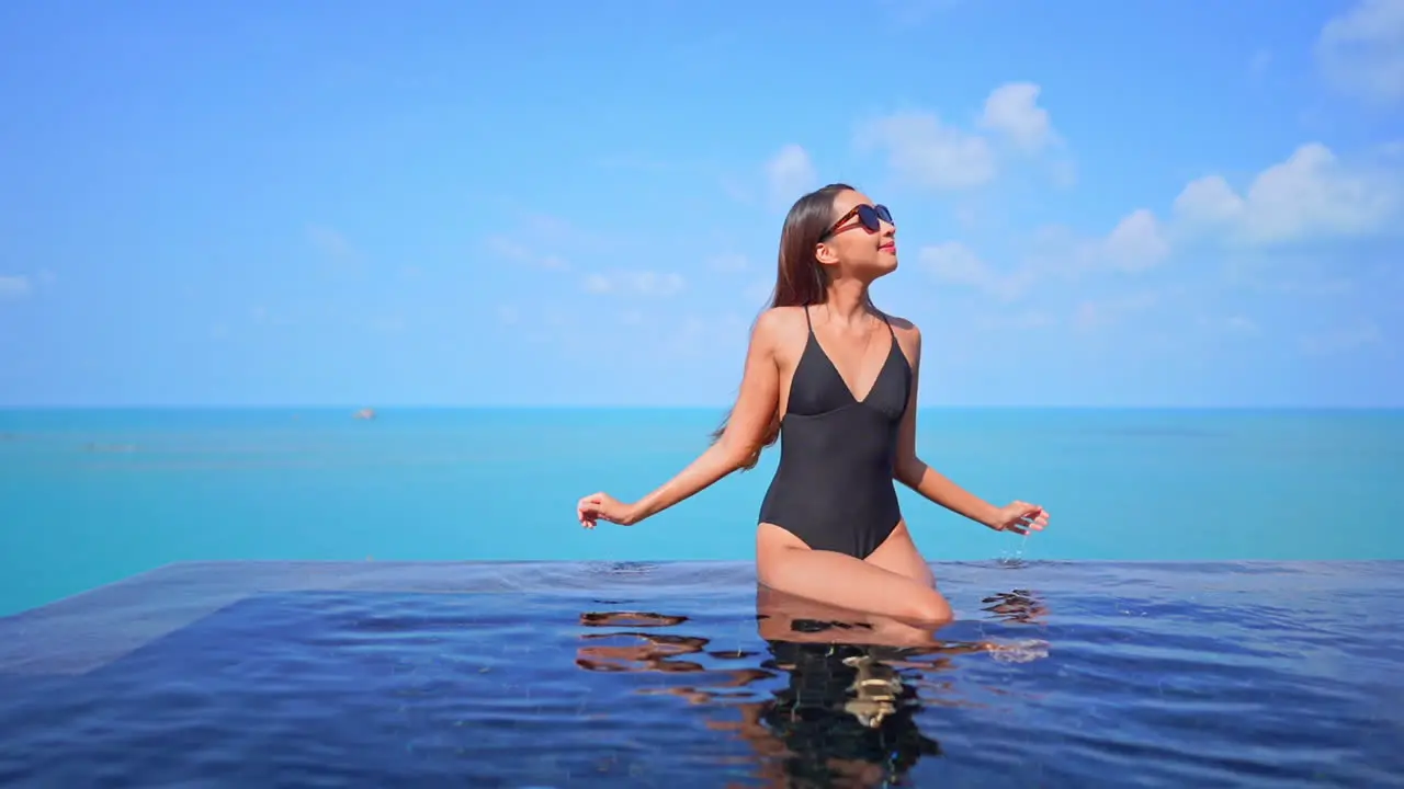 Pretty woman in swimwear sitting on rooftop infinity pool border and raising hands up with a stunning view of endless turquoise seascape on horizon in Phuket Thailand static copy space