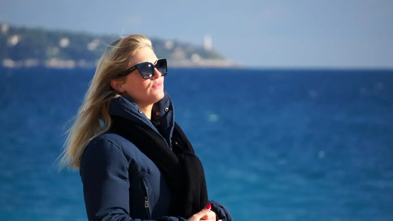 A young woman waves her blond hair in the wind the blue sea in soft focus behind her on a sunny windy day SLOMO