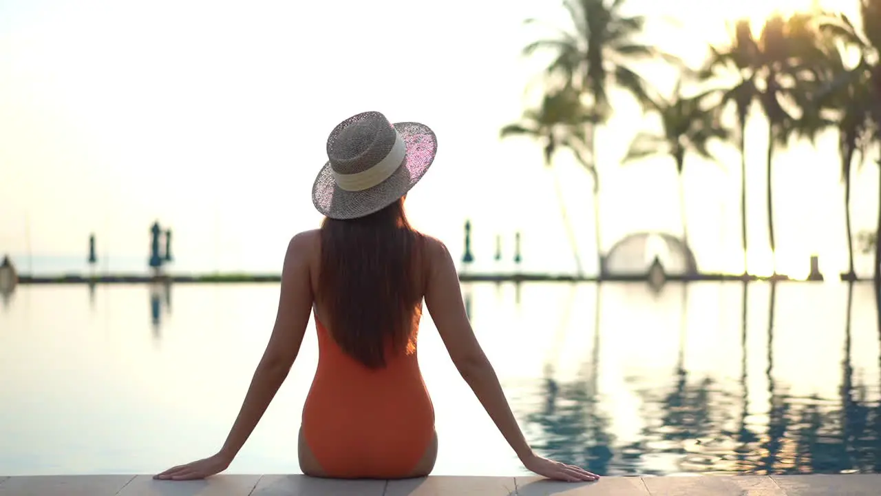 Back view of the woman in an orange swimsuit sitting on the edge of the swimming pool at an exotic hotel in Hawaii on sunset