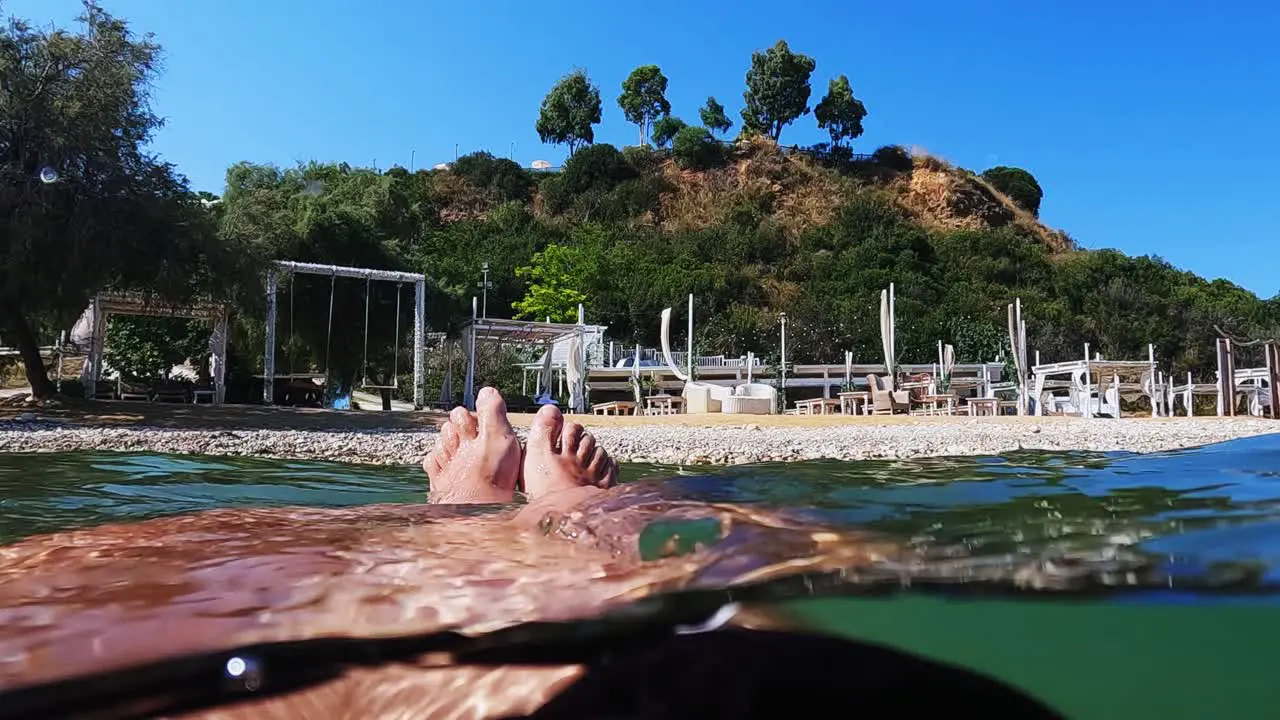 Personal perspective of male legs and feet relaxing while floating on sea water with swing on beach in background