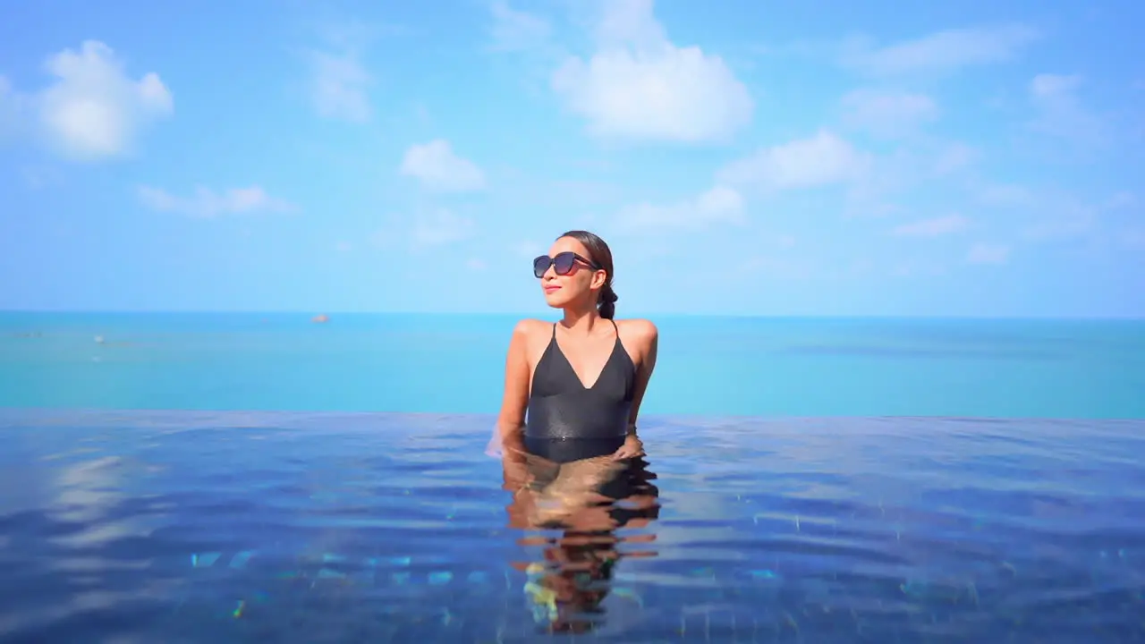 Fit Woman Model in Black Swimming Suit Leaning Arms on the Edge of Infinity Pool on Turquoise Seascape Background Wearing Sunglasses on a Sunny Day Slow Motion