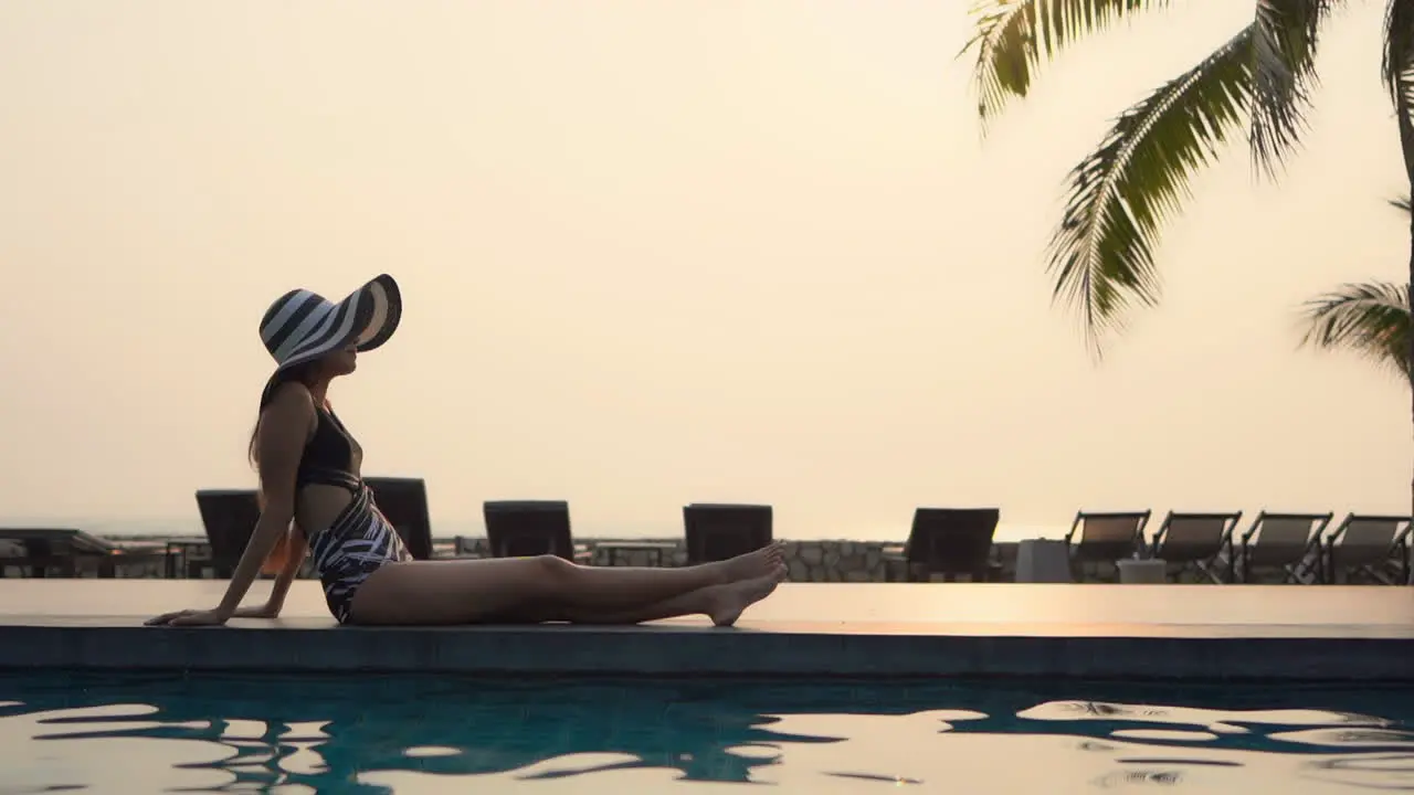 Slow-motion on the silhouette of Asian model woman sitting at the edge of the swimming pool on sunset in Thailand resort side view