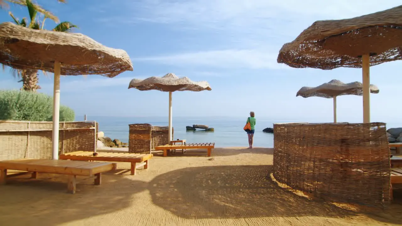 Steadicam Shot Woman Standing On The Beach Near The Parasols And The Palm Trees Looking At The Sea