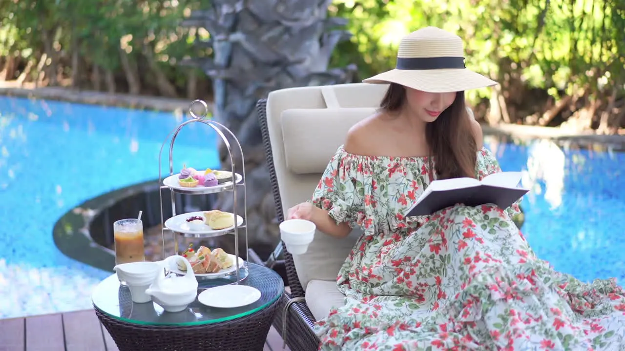 Elegant woman with hat and floral dress sips tea while reading book poolside