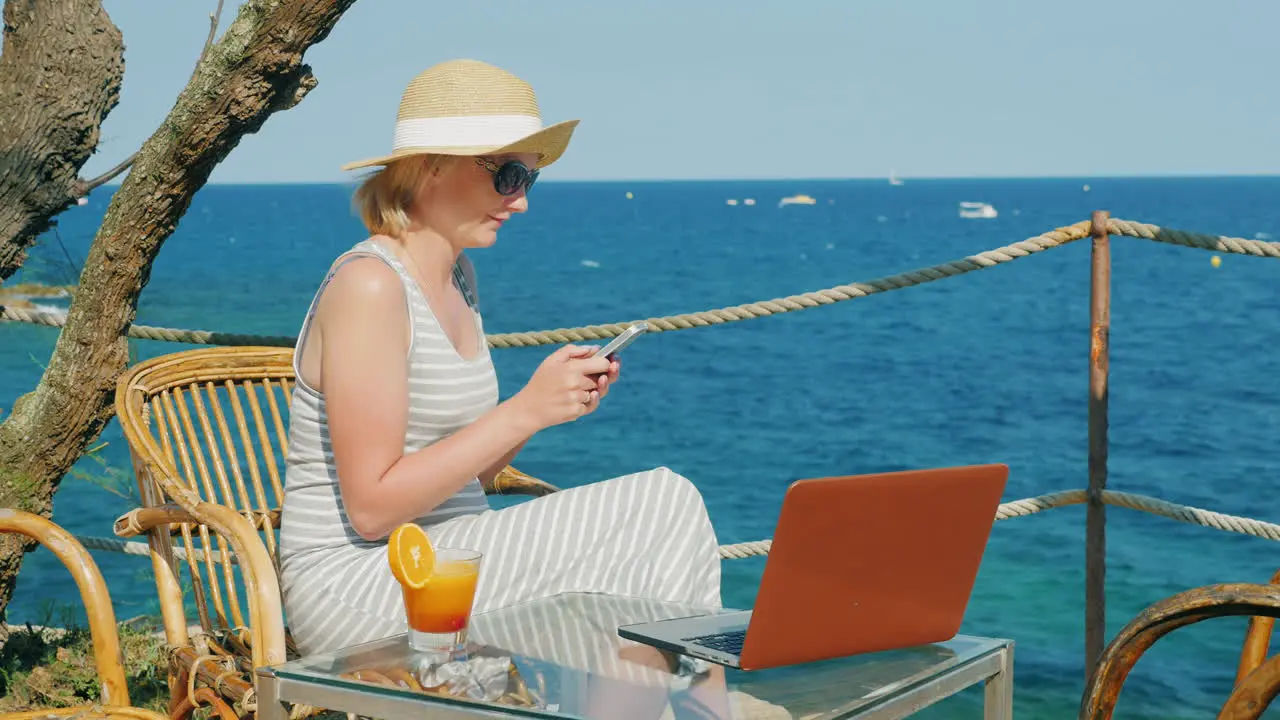 Woman Tourist Relaxing In A Cafe Overlooking The Sea Enjoys A Smartphone Standing Next To A Laptop