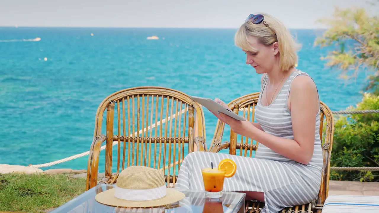 Young Woman Enjoys The Tablet Sitting On The Summer Terrace Cafe Overlooking The Sea