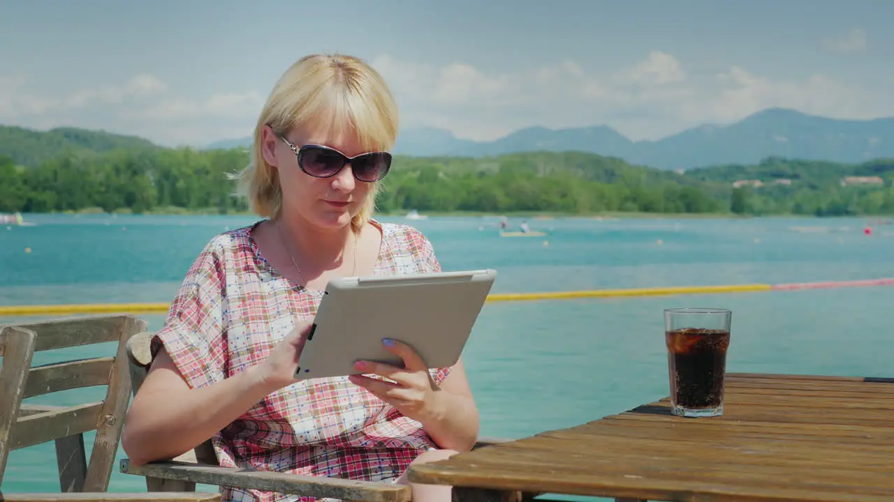 Technology On Vacation Woman With Tablet Sitting At A Table In A Cafe On The Background Of The Lake 