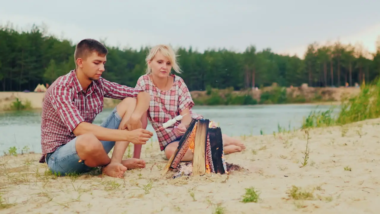 Two Friends Are Heated By The Fire Roast Marshmallows On Sticks Against The Backdrop Of The River An