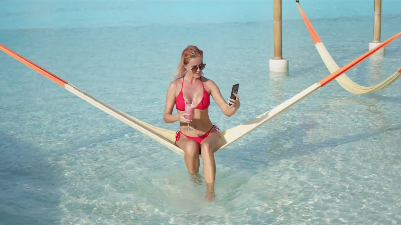 Cheerful woman with cocktail taking selfie on hammock