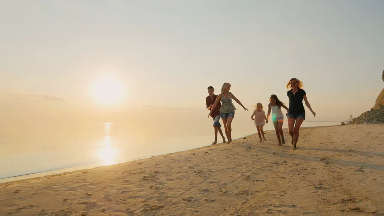 Group Of Carefree Children Of Different Ages And Adults Have Fun Running On The Beach At Sunset