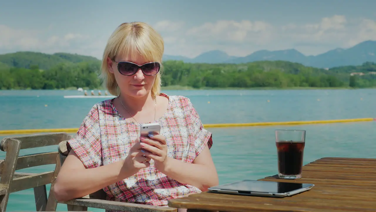 Business Woman Uses A Smartphone On Vacation Sitting At A Table In A Cafe Near The Lake On A Backgro