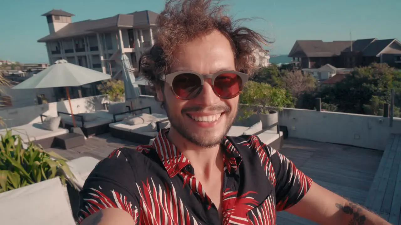 Young Man In Sunglasses Waving Hands To The Camera In The Foreground Terrace With Pool And Hammocks In The Background