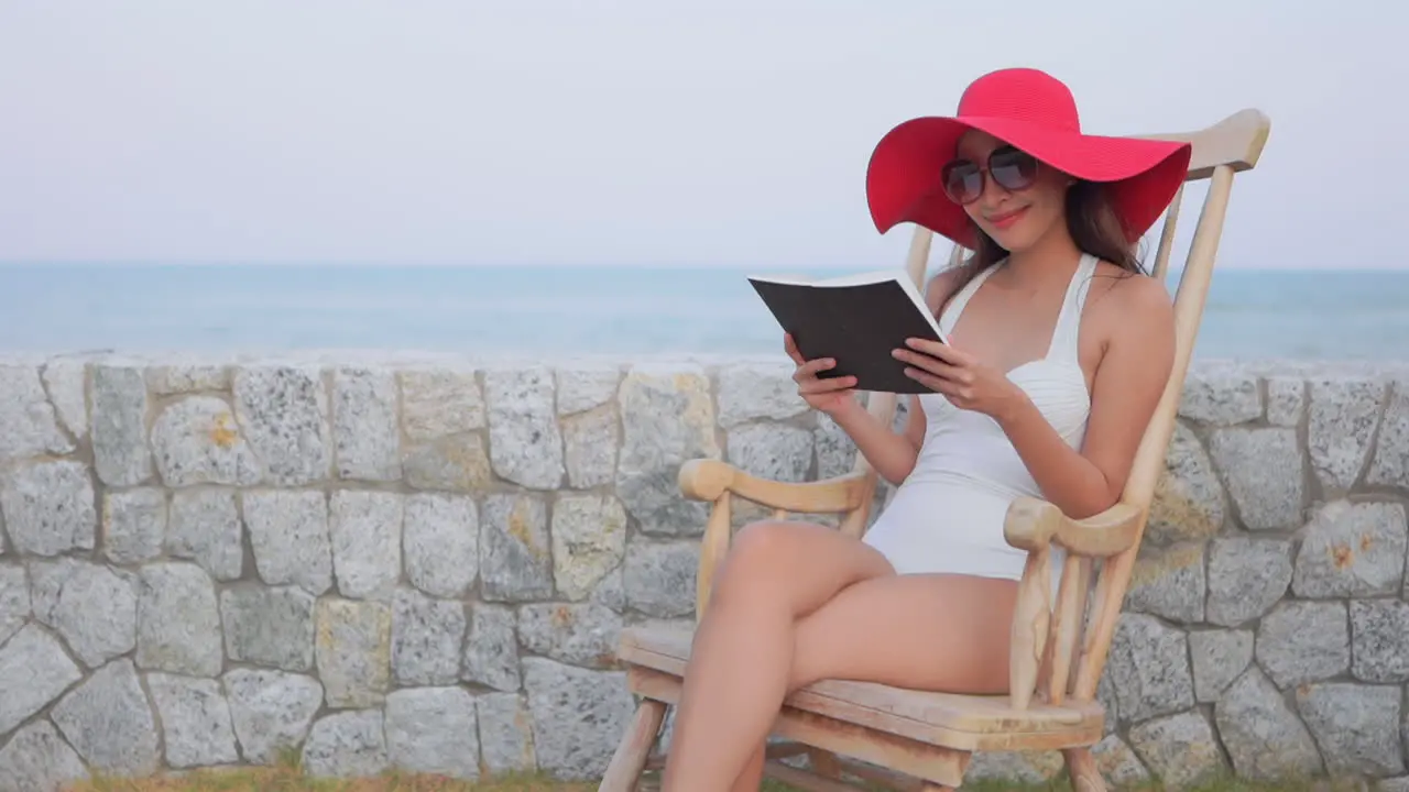 Asian woman with a beautiful smile is reading a magazine sitting on a wooden chair near the beach wearing white monokini and a big red hat