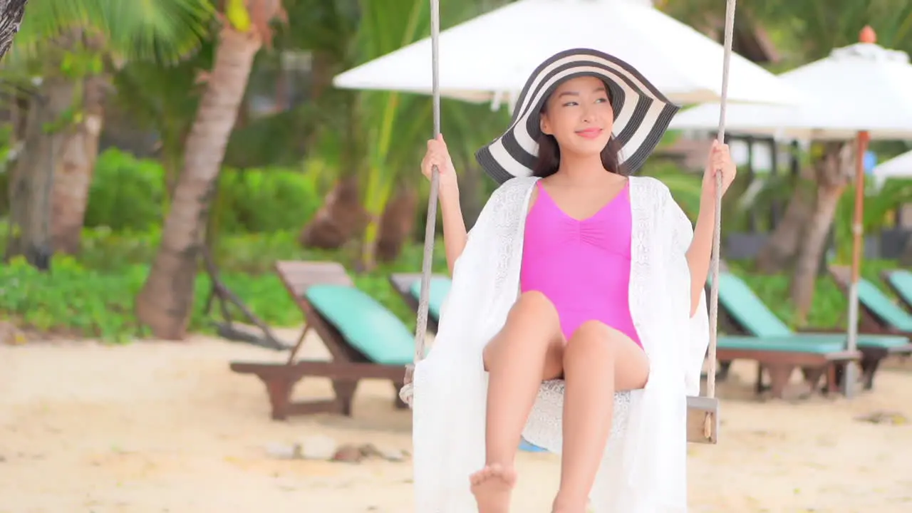 Asian tourist smiling while swinging on swing at tropical beach on sunny day wearing pink swimsuit and big striped hat