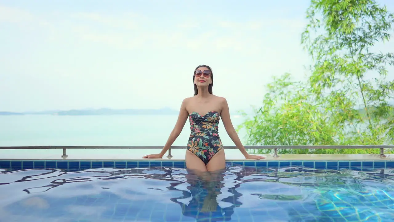 With the ocean horizon in the background an attractive young woman leans along the edge of a resort pool enjoying the sun