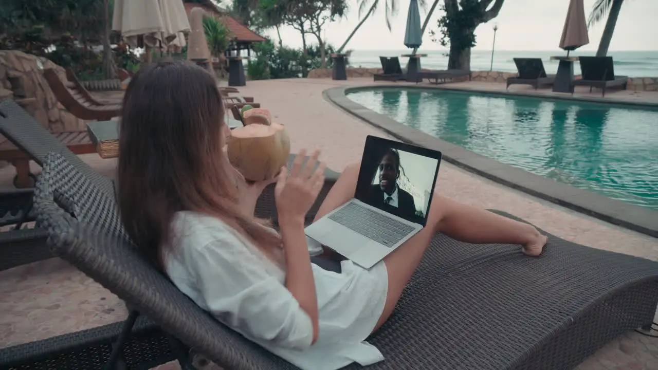 Woman Wearing White Shirt And Drinking From A Coconut On Video Call With An Coworker While Sitting In The Hammock In Front Of The Pool In Resort