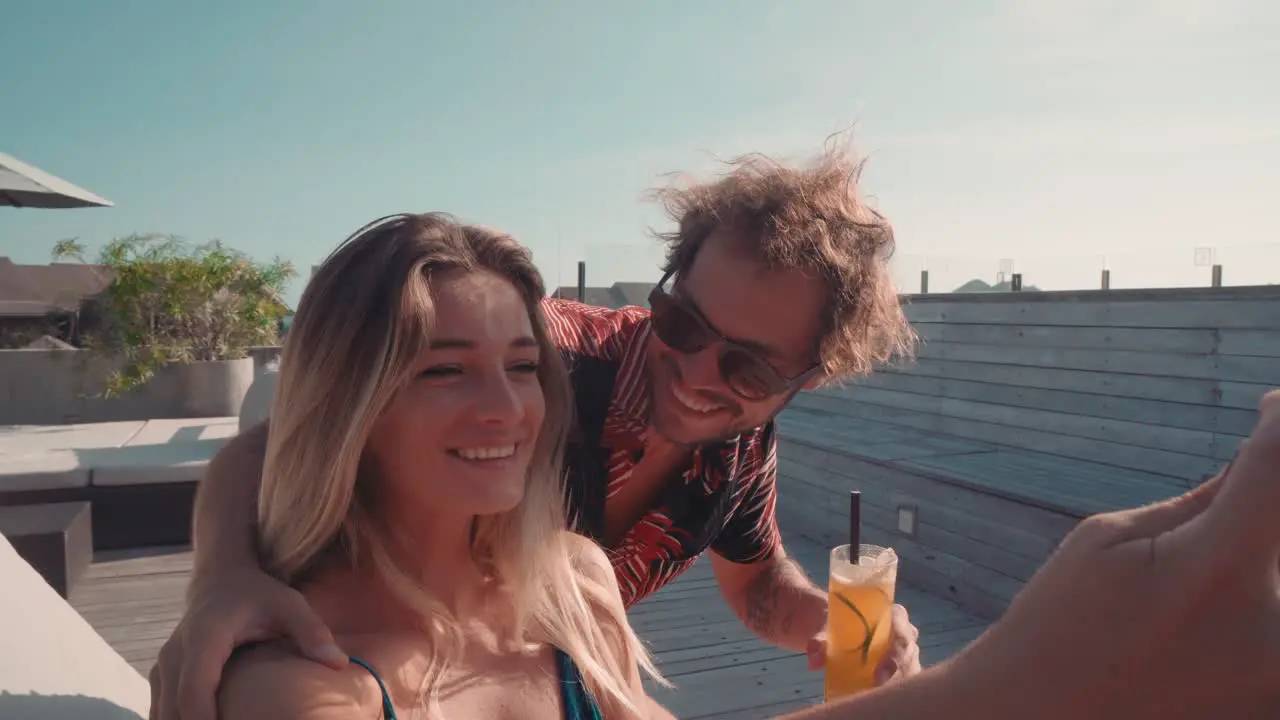 Young Man Holding A Drink And Taking A Selfie With A Blonde Young Woman Sitting In A Hammock In A Resort 1