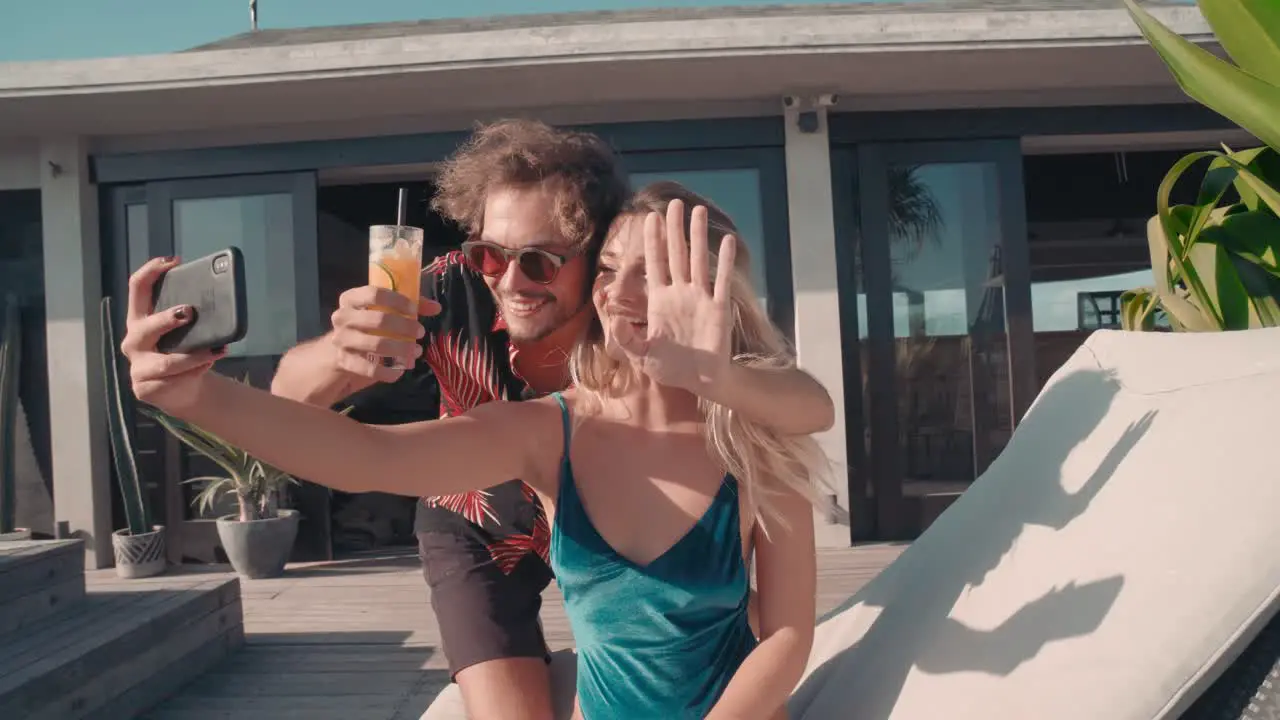 Young Man Goes Out To The Terrace Holding A Drink Taking A Selfie With A Blonde Young Woman Sitting In A Hammock