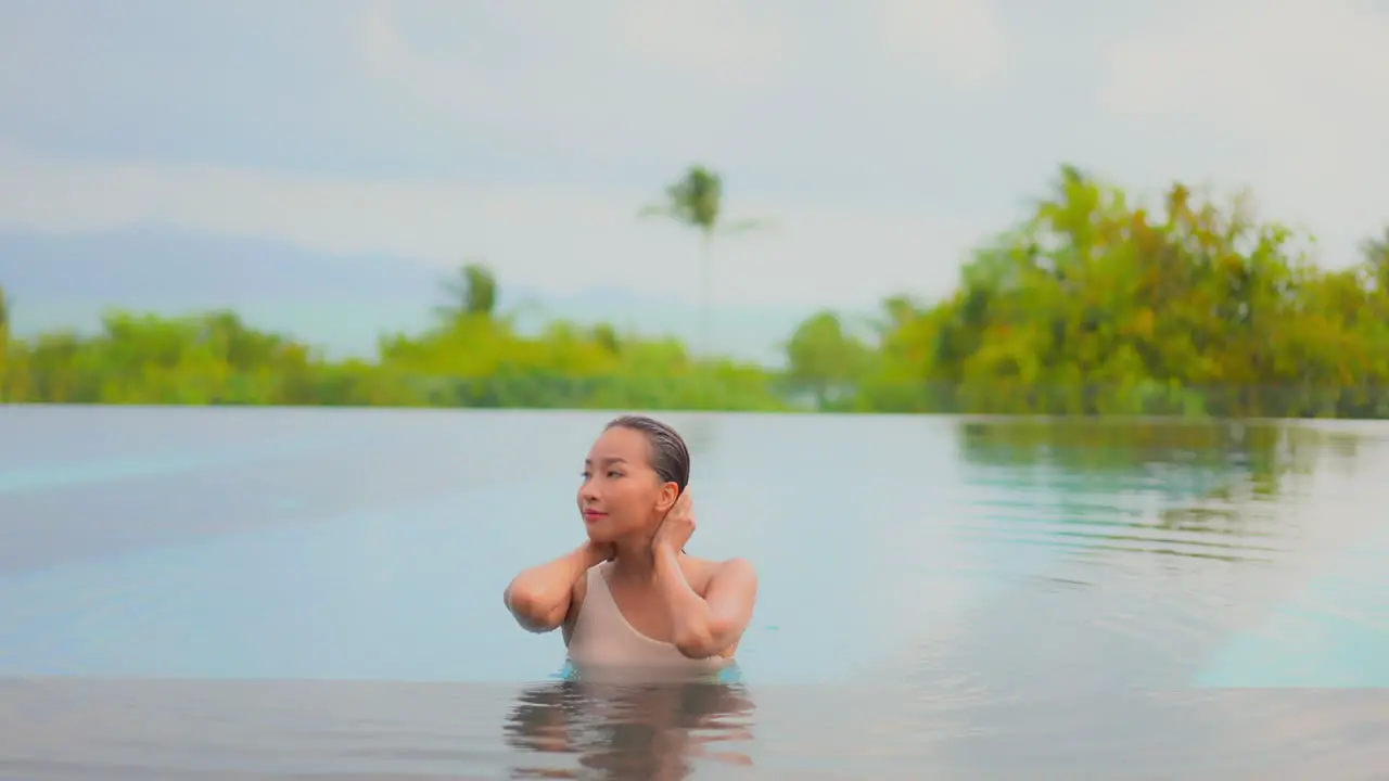 Attractive young woman touches her wet hair inside the swimming pool water of an exotic Bali hotel with closed eyes on a sunny day portrait slow motion