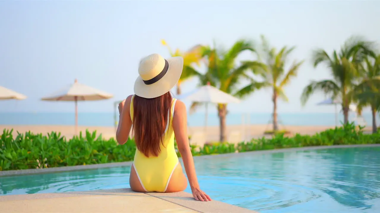 Backside view of the woman sitting on edge of swimming pool at exotic hotel in Hawaii near the beach in yellow monokini and white hat and hitting water with her legs beach umbrellas and Palms