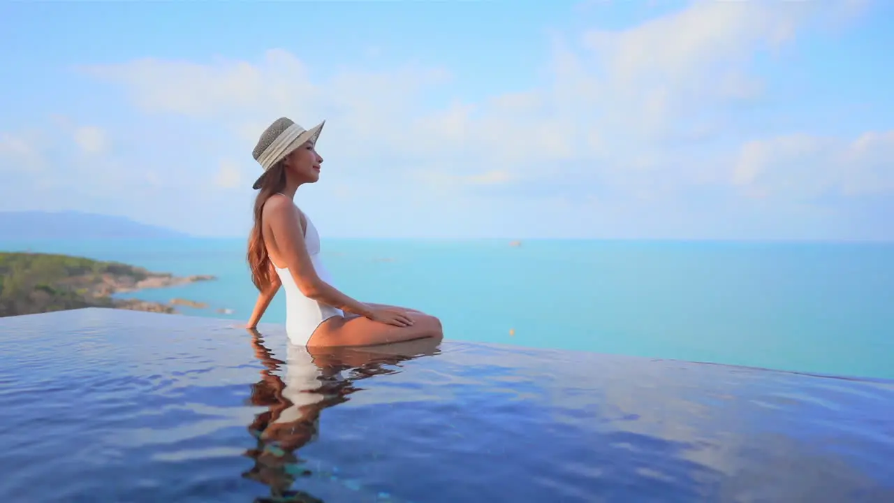 A fit young healthy woman sits on the edge of a resort pool while looking out over the ocean horizon