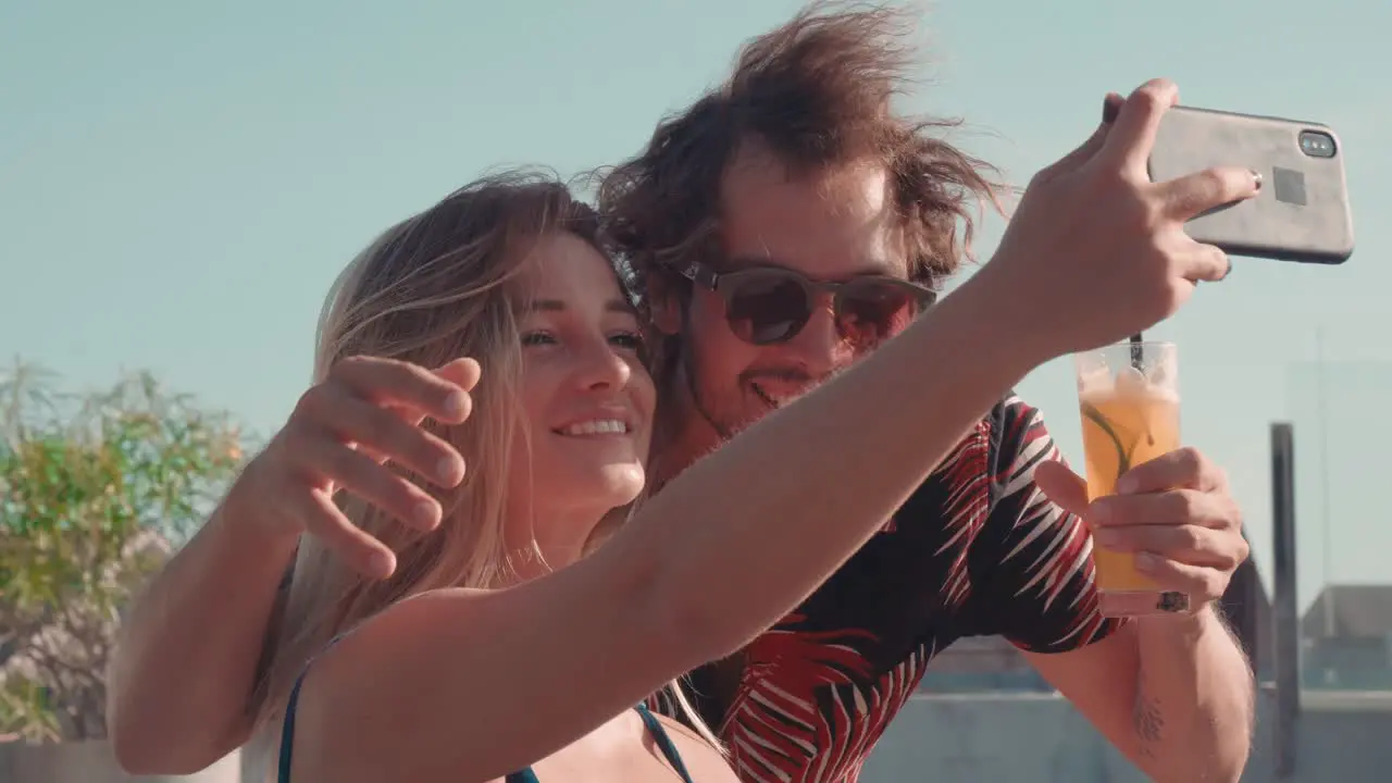 Young Man Holding A Drink And Taking A Selfie With A Blonde Young Woman Sitting In A Hammock In A Resort