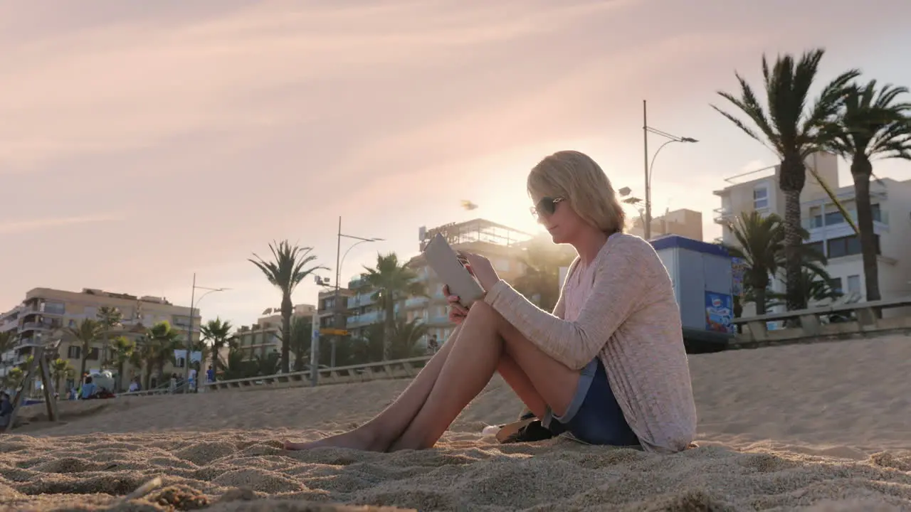 A young woman uses a tablet sitting on the beach against the backdrop of palm trees in Spain