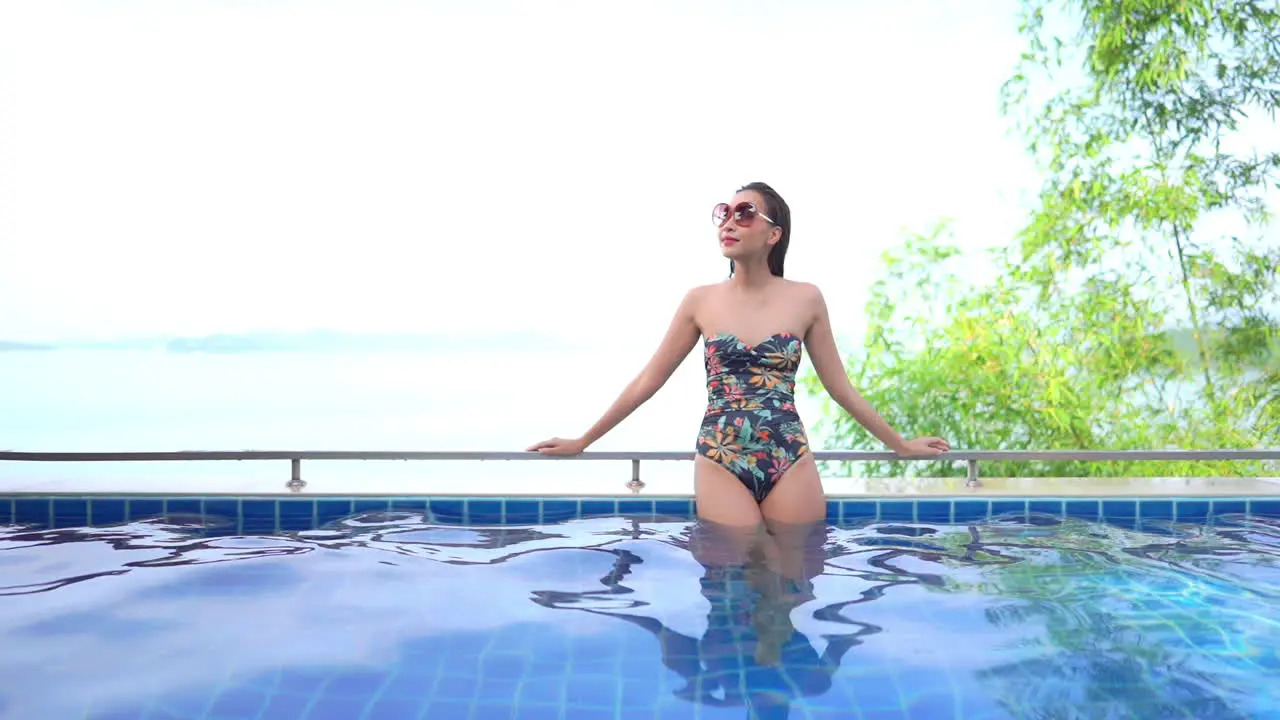 Asian woman resting leaning on the swimming pool handrail with seascape behind during summer vacation