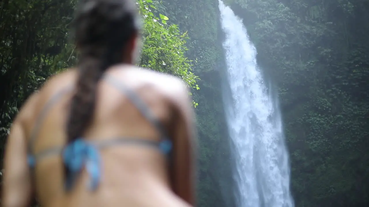 Slow motion panning shot of a girl in a blue bikini sitting in front of a gushing NungNung Waterfall in Bali Indonesia