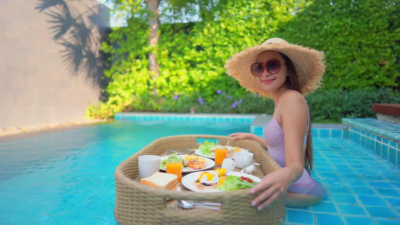 Side view of an asian tourist sitting in the swimming pool with a floating breakfast tray full of delicious and healthy food