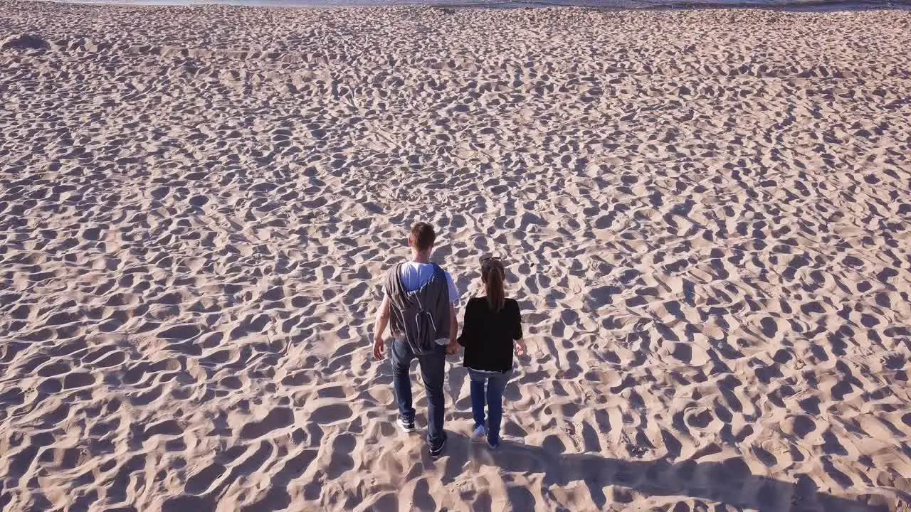 Aerial Shot Of Young Couple Walking On Sandy Beach Holding Hands And Running Towards Water