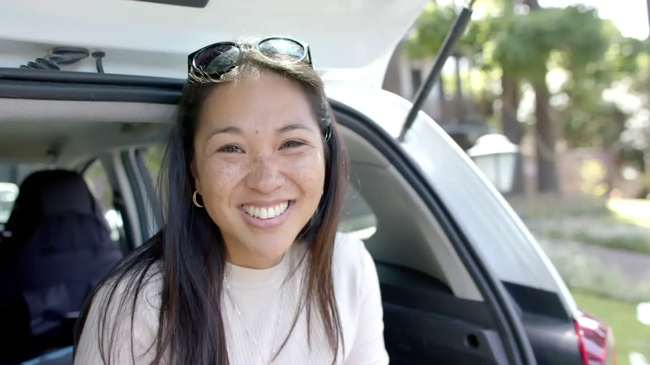 Happy asian woman with sunglasses sitting in car trunk and smiling on sunny day slow motion