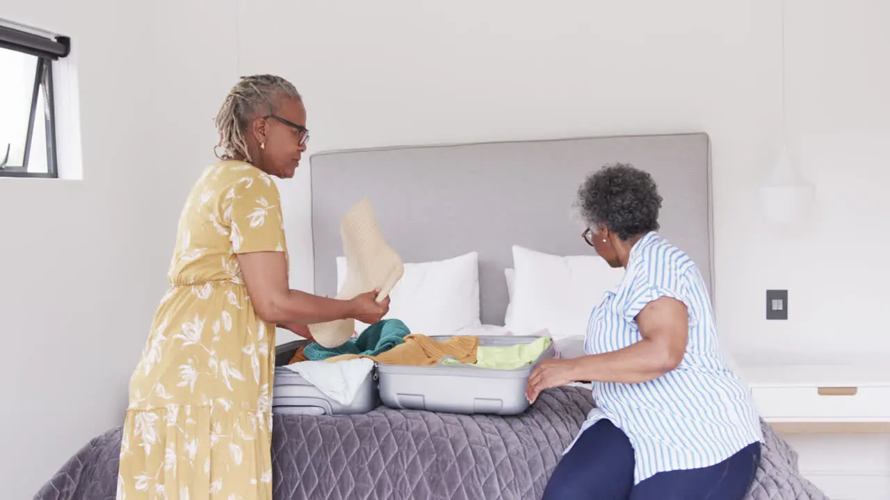 Happy senior african american female friends unpacking suitcase on bed at holiday home slow motion