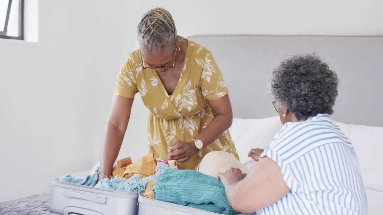 Happy senior african american female friends packing suitcase on bed at holiday home slow motion