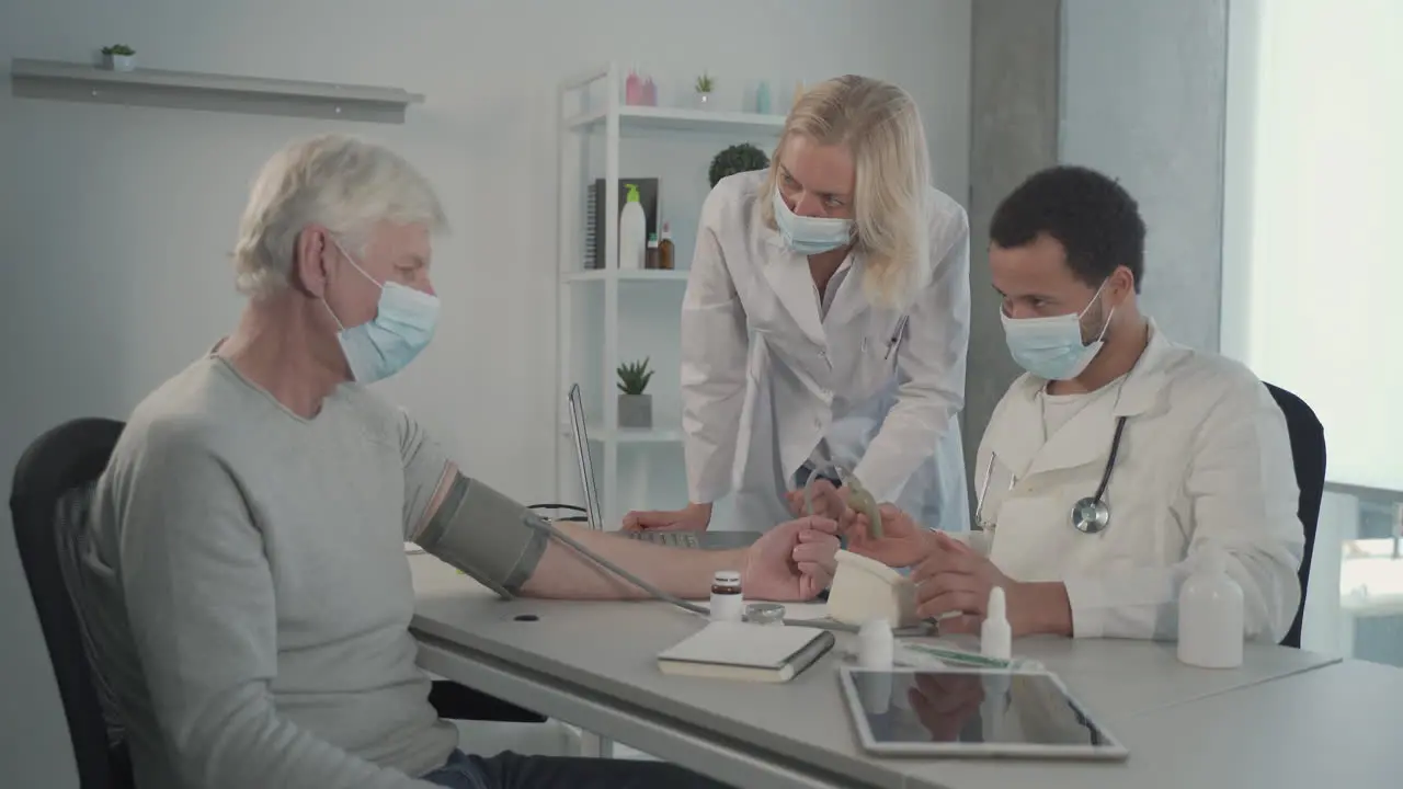 A Young Black Doctor In A Facemask Takes Blood Pressure To A Grey Haired Man Under The Gaze Of A Female Blonde Doctor