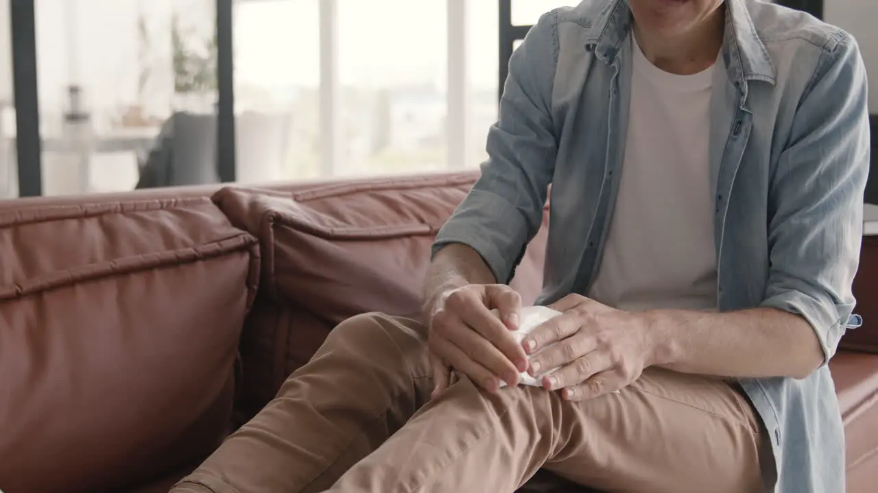 Middle Aged Man Trying To Relieve Knee Pain With An Ice Pack While Sitting On Sofa At Home