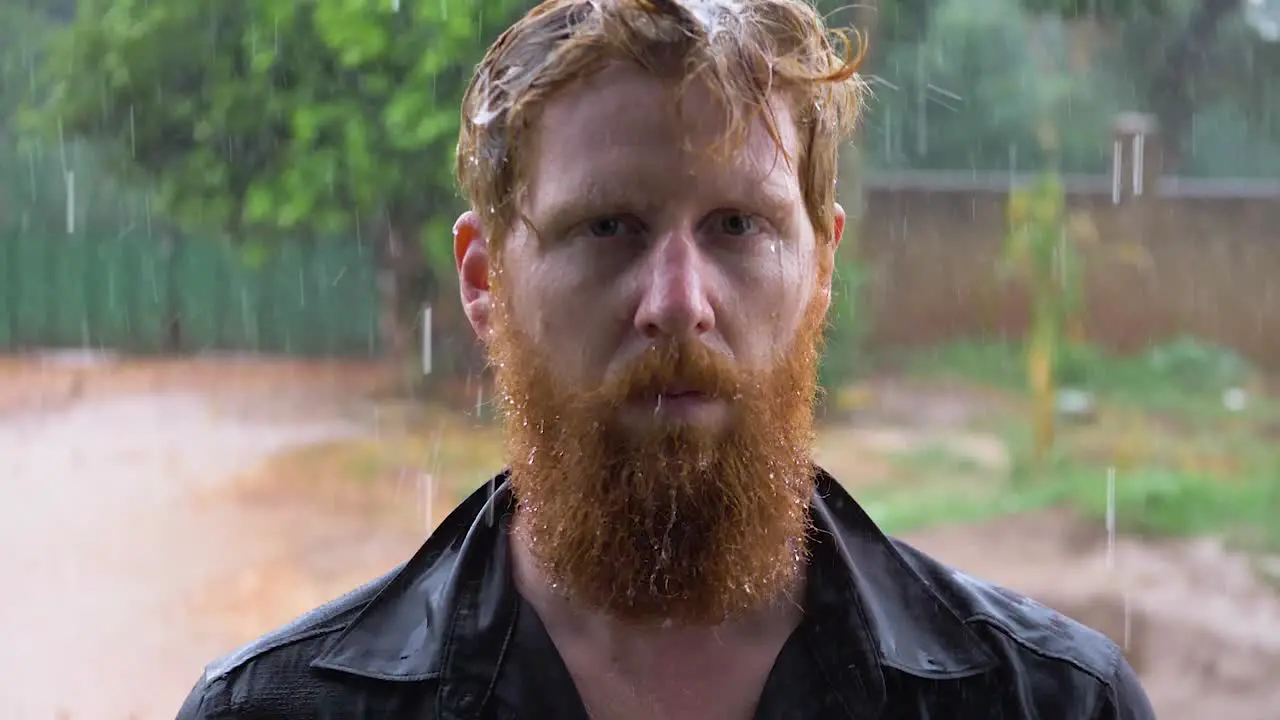 Slow motion portrait shot of a bearded ginger man standing in the pouring rain and getting soaked while looking into the camera