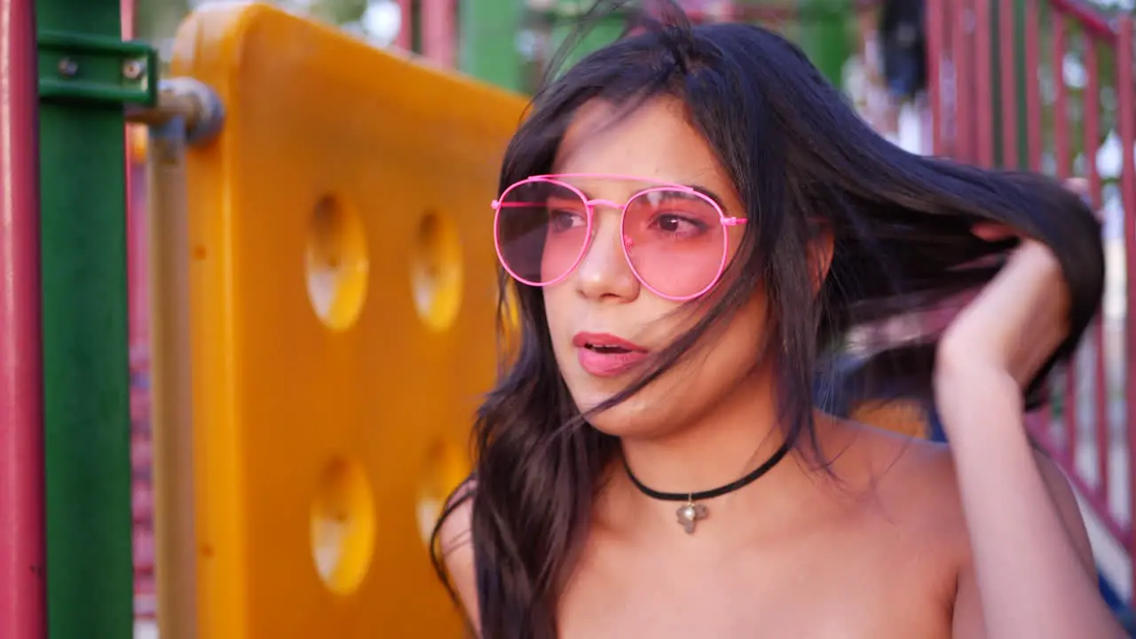 A young hispanic woman hipster wearing vintage fashion clothing and retro pink aviator sunglasses in a park playground SLOW MOTION